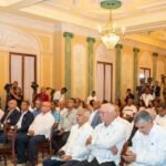 Arroceros rechazan las medidas del Gobierno frente al arroz de EE. UU. Asistentes sentados en un salón con candelabros, columnas verdes y cámaras de prensa al fondo durante un acto oficial