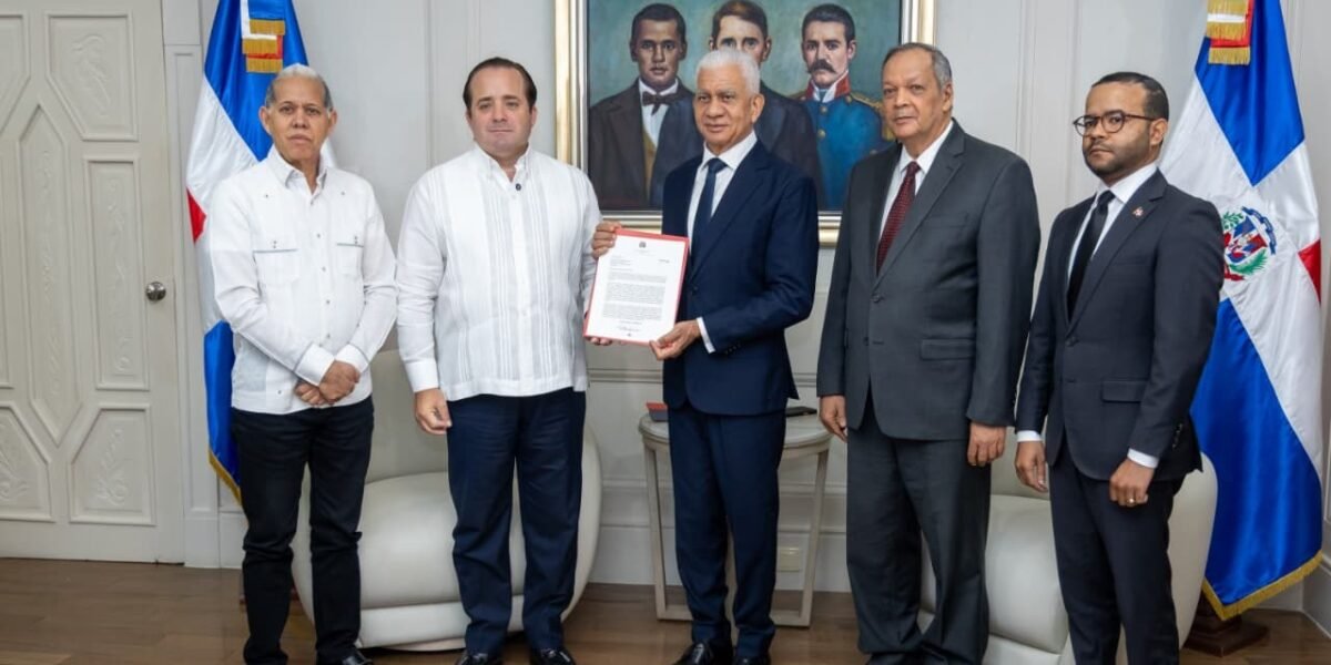 Cinco funcionarios posan de pie entre banderas dominicanas mientras dos de ellos sostienen un documento
