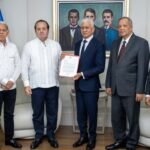 Paliza entrega al Senado el Plan Nacional de Ordenamiento Territorial Cinco funcionarios posan de pie entre banderas dominicanas mientras dos de ellos sostienen un documento
