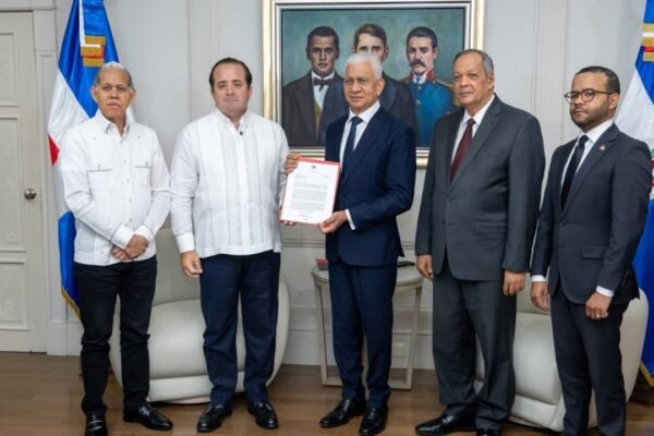 Cinco funcionarios posan de pie entre banderas dominicanas mientras dos de ellos sostienen un documento