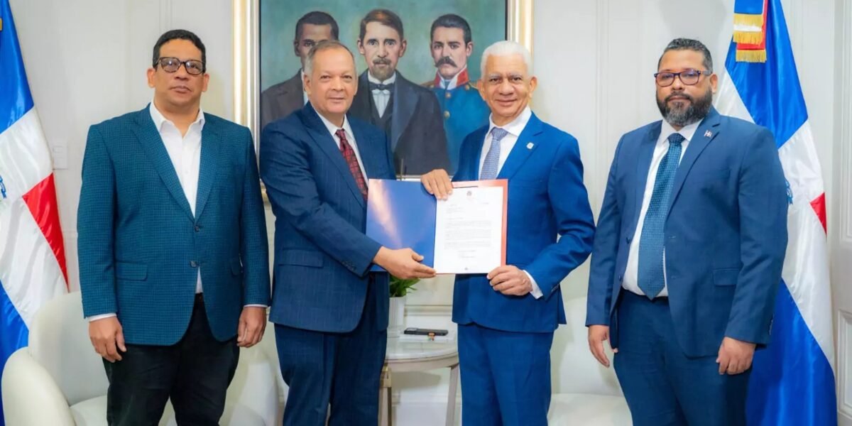 Cuatro hombres de traje posan entre banderas dominicanas; dos de ellos sostienen una carpeta azul con un documento oficial, frente a un cuadro de los padres de la patria