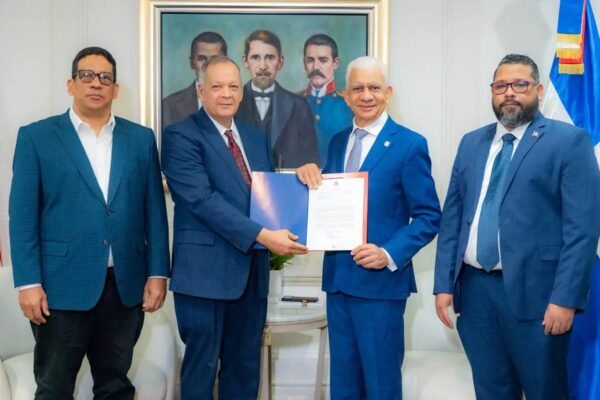 Cuatro hombres de traje posan entre banderas dominicanas; dos de ellos sostienen una carpeta azul con un documento oficial, frente a un cuadro de los padres de la patria