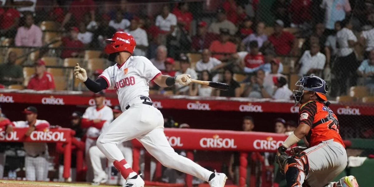 Un bateador de los Leones del Escogido, con casco rojo y uniforme blanco, sale a correr tras hacer contacto; a la derecha, el receptor de los Toros del Este con uniforme anaranjado