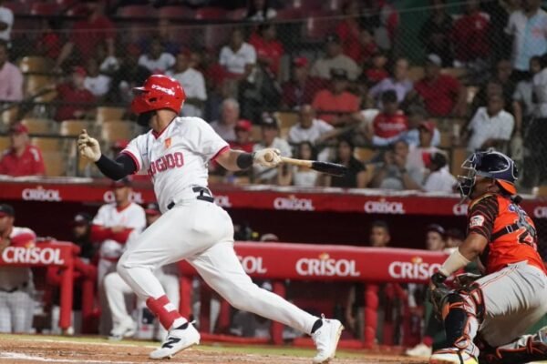 Un bateador de los Leones del Escogido, con casco rojo y uniforme blanco, sale a correr tras hacer contacto; a la derecha, el receptor de los Toros del Este con uniforme anaranjado