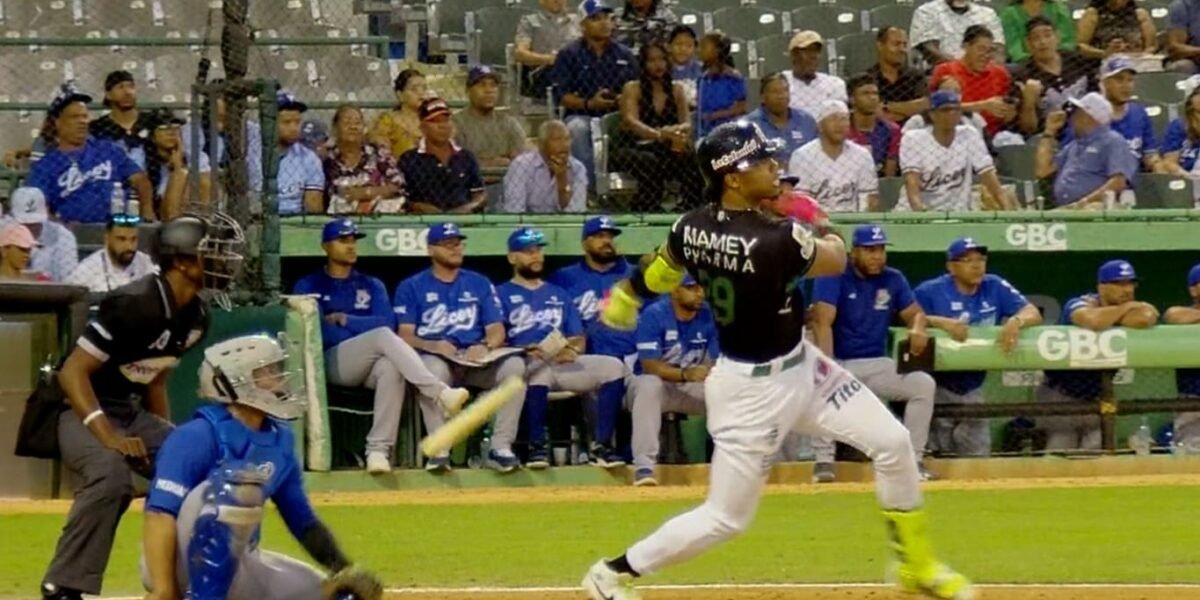 Un bateador con uniforme negro y verde de las Estrellas Orientales sigue la pelota tras su swing; detrás, el receptor y el umpire, y al fondo el dugout del Licey y el público en las gradas.
