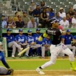 Un bateador con uniforme negro y verde de las Estrellas Orientales sigue la pelota tras su swing; detrás, el receptor y el umpire, y al fondo el dugout del Licey y el público en las gradas.