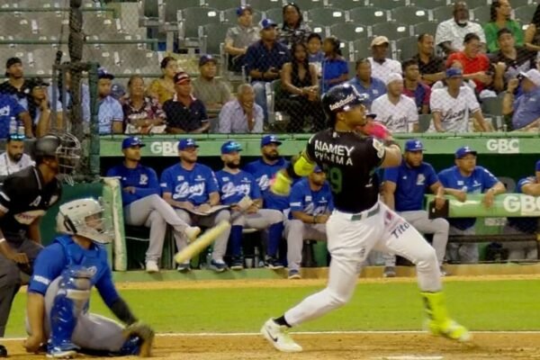Un bateador con uniforme negro y verde de las Estrellas Orientales sigue la pelota tras su swing; detrás, el receptor y el umpire, y al fondo el dugout del Licey y el público en las gradas.