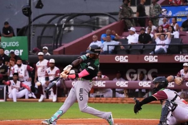 Un bateador de las Estrellas Orientales, con uniforme gris y verde, hace swing ante el receptor de los Gigantes del Cibao, de uniforme blanco y vinotinto, con el dugout y el público al fondo