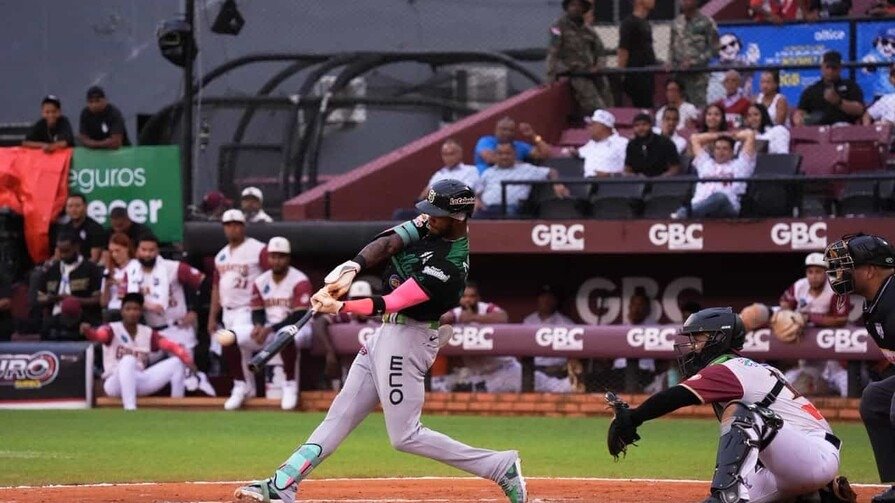 Un bateador de las Estrellas Orientales, con uniforme gris y verde, hace swing ante el receptor de los Gigantes del Cibao, de uniforme blanco y vinotinto, con el dugout y el público al fondo