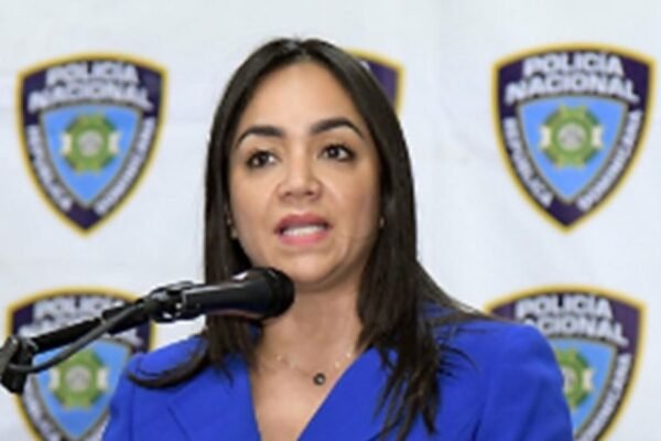 Faride Raful habla ante un micrófono, con chaqueta azul, frente a un fondo con escudos de la Policía Nacional.