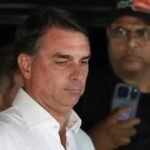 Flávio Bolsonaro llega a Miami pese a la orden de entregar sus pasaportes Flávio Bolsonaro, con camisa blanca, mira hacia abajo con gesto serio; detrás, un hombre con gorra y lentes sostiene un teléfono