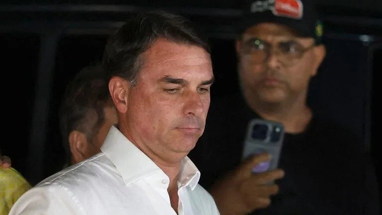 Flávio Bolsonaro, con camisa blanca, mira hacia abajo con gesto serio; detrás, un hombre con gorra y lentes sostiene un teléfono