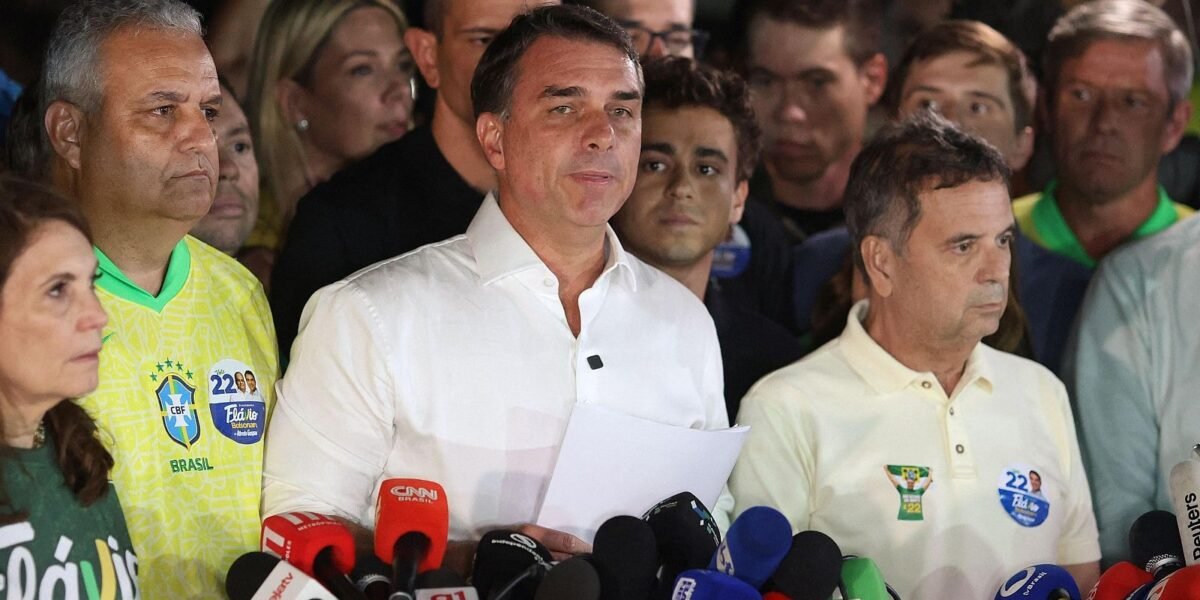Flávio Bolsonaro, con camisa blanca y unos papeles en la mano, habla ante varios micrófonos rodeado de simpatizantes