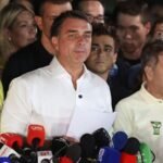 Flávio Bolsonaro, con camisa blanca y unos papeles en la mano, habla ante varios micrófonos rodeado de simpatizantes