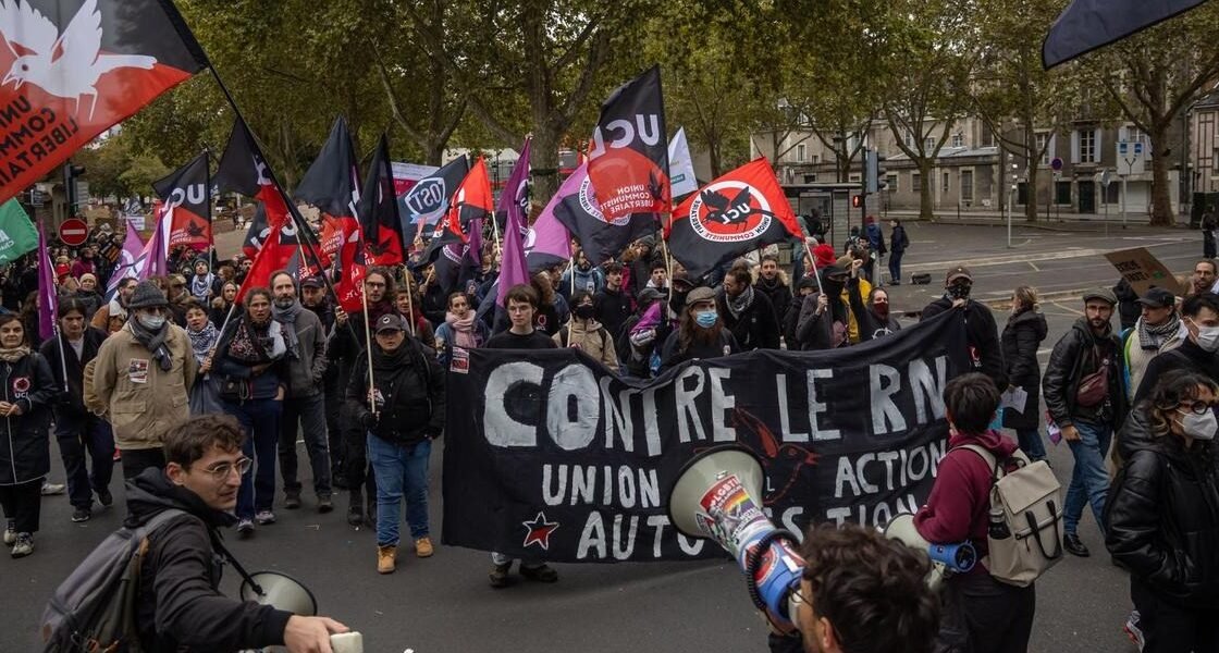 Manifestantes con banderas rojas y negras y una pancarta que dice «Contre le RN» marchan por una avenida arbolada; en primer plano, dos personas con megáfonos