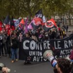 Manifestantes con banderas rojas y negras y una pancarta que dice «Contre le RN» marchan por una avenida arbolada; en primer plano, dos personas con megáfonos