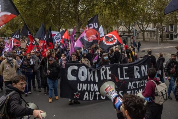 Manifestantes con banderas rojas y negras y una pancarta que dice «Contre le RN» marchan por una avenida arbolada; en primer plano, dos personas con megáfonos
