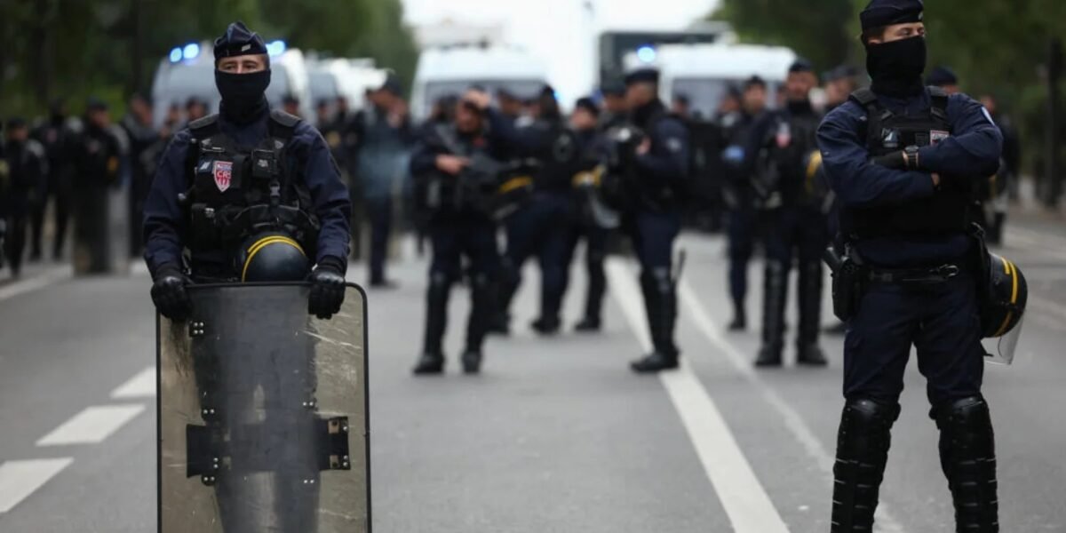Agentes antidisturbios franceses con pasamontañas, chalecos y escudo forman un cordón en una avenida, con furgonetas policiales al fondo