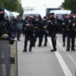 Agentes antidisturbios franceses con pasamontañas, chalecos y escudo forman un cordón en una avenida, con furgonetas policiales al fondo