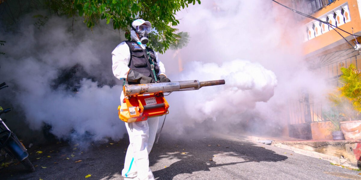 Trabajador con traje protector blanco, mascarilla antigás y gafas fumiga con una termonebulizadora en una calle residencial cubierta de humo.