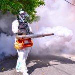 Trabajador con traje protector blanco, mascarilla antigás y gafas fumiga con una termonebulizadora en una calle residencial cubierta de humo.