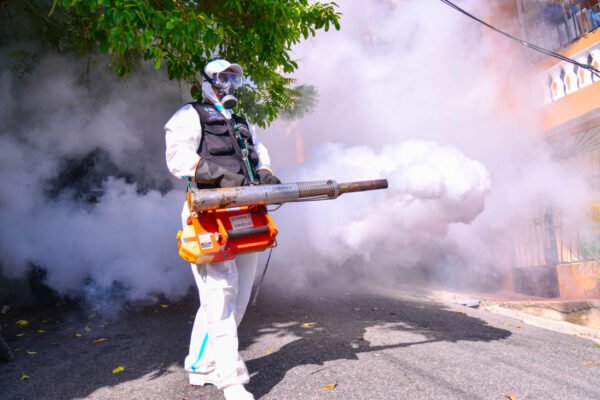 Trabajador con traje protector blanco, mascarilla antigás y gafas fumiga con una termonebulizadora en una calle residencial cubierta de humo.