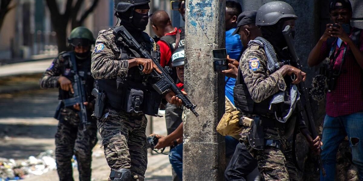 Agentes con uniforme de camuflaje, casco y fusiles avanzan por una calle mientras reporteros gráficos toman imágenes
