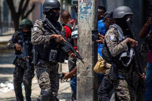 Agentes con uniforme de camuflaje, casco y fusiles avanzan por una calle mientras reporteros gráficos toman imágenes