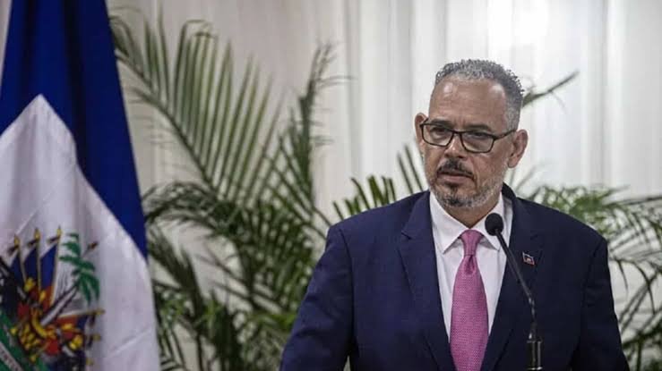 Un hombre con gafas y traje oscuro habla ante un micrófono junto a una bandera de Haití