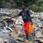 Un joven camina con un bidón amarillo entre la basura acumulada en un cauce