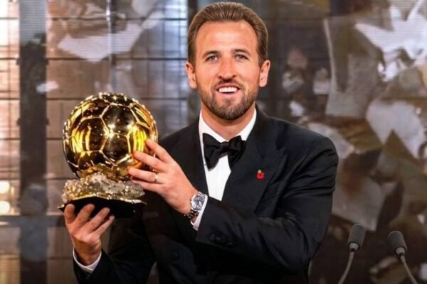 Harry Kane, con esmoquin y pajarita negra, sonríe mientras sostiene el trofeo del Balón de Oro junto a dos micrófonos