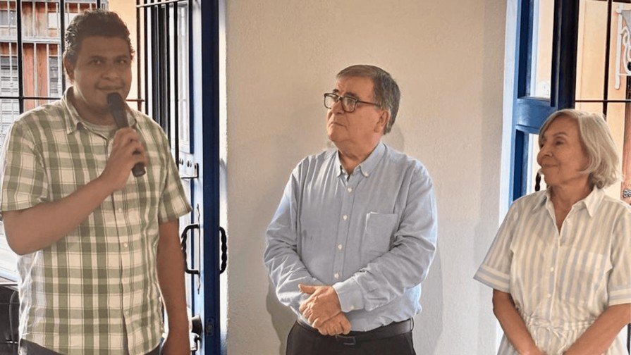 Un hombre con camisa a cuadros habla por un micrófono junto a un hombre con lentes y camisa azul y una mujer de cabello canoso, en una sala con rejas azules.