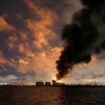 Columna de humo negro y llamas sobre los tanques de una terminal petrolera junto al agua, de noche