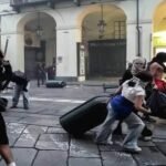 Manifestantes encapuchados arrastran maletas y objetos bajo los soportales de una calle comercial italiana durante disturbios