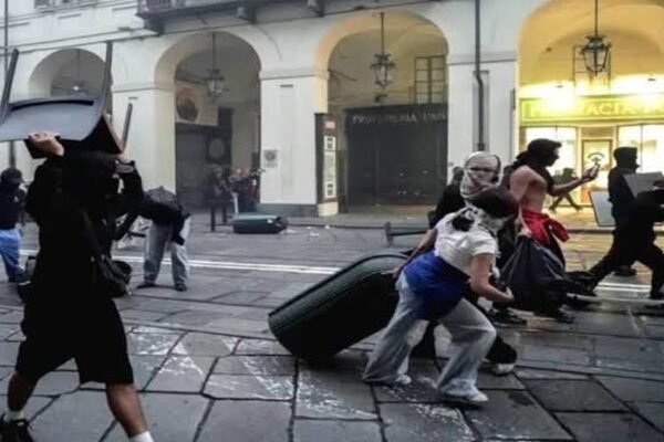 Manifestantes encapuchados arrastran maletas y objetos bajo los soportales de una calle comercial italiana durante disturbios