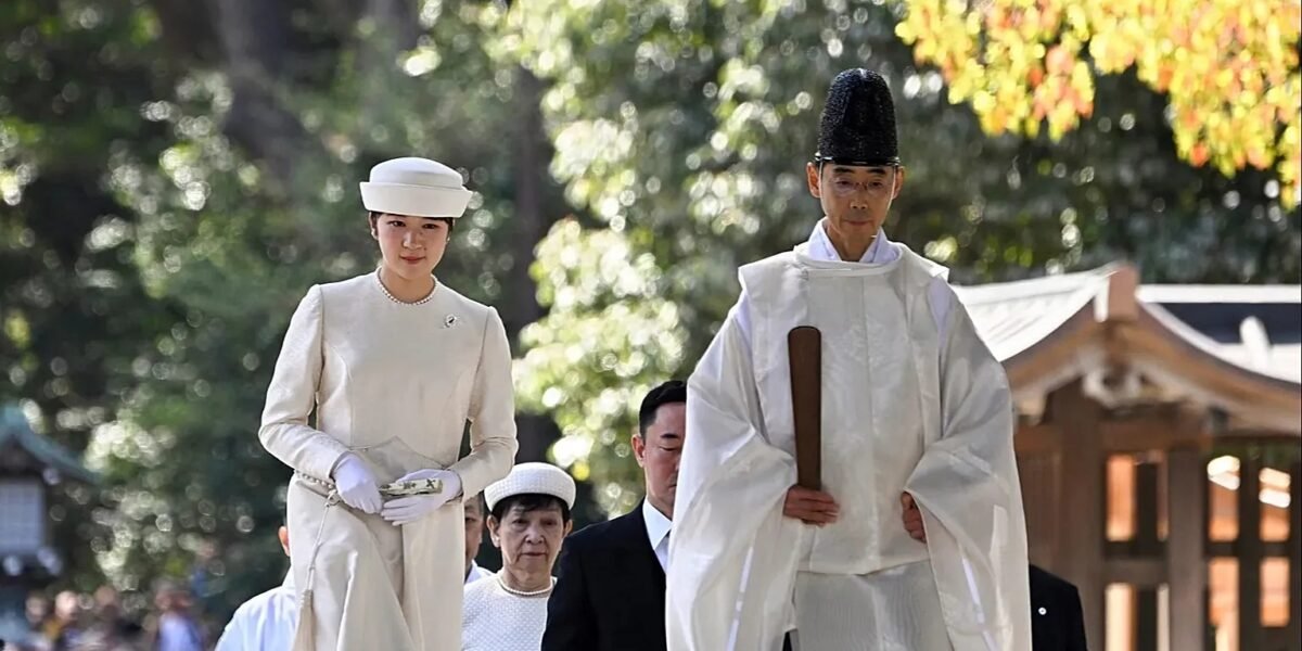 Una joven con vestido y sombrero blancos camina detrás de un sacerdote sintoísta con túnica blanca y tocado negro en el recinto arbolado de un santuario japonés