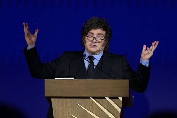 Javier Milei, con gafas y traje oscuro, habla con los brazos levantados detrás de un atril de madera ante un fondo azul