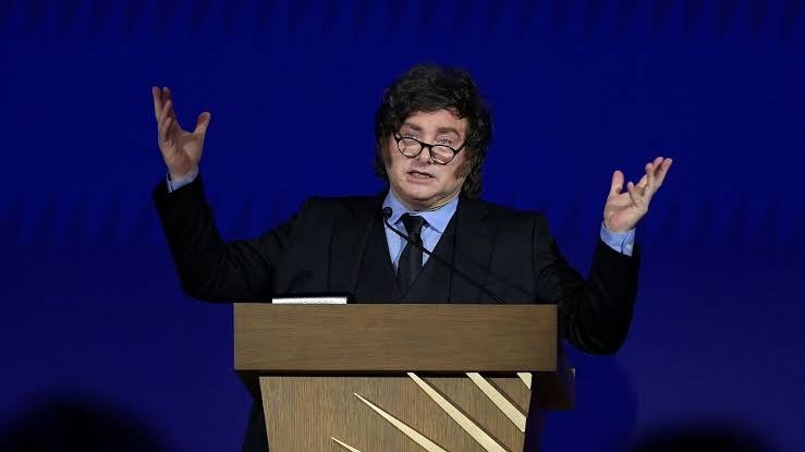 Javier Milei, con gafas y traje oscuro, habla con los brazos levantados detrás de un atril de madera ante un fondo azul