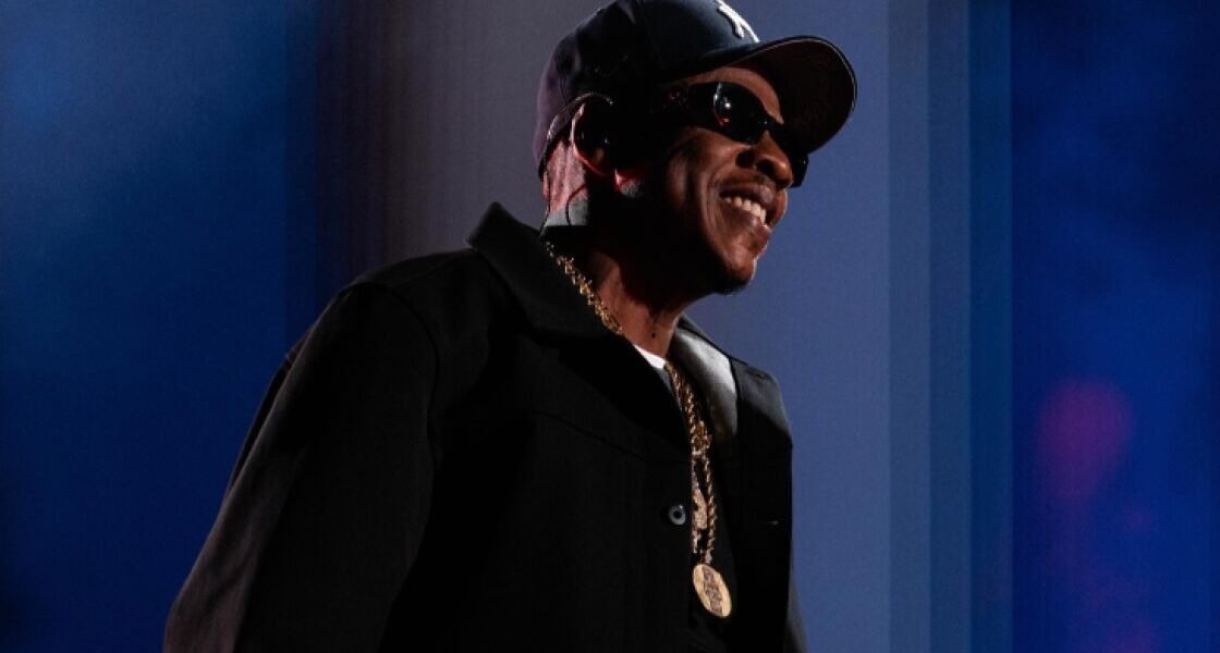 Jay-Z sonríe en un escenario con gorra oscura, gafas de sol, chaqueta negra y una cadena dorada, ante un fondo azul.