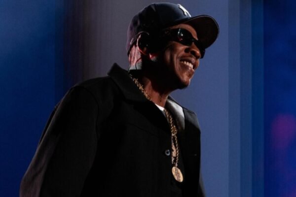 Jay-Z sonríe en un escenario con gorra oscura, gafas de sol, chaqueta negra y una cadena dorada, ante un fondo azul.