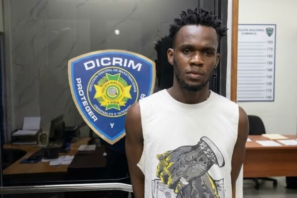 Un hombre joven con camiseta blanca sin mangas posa frente a una puerta de vidrio con el escudo de la Dirección Central de Investigaciones (Dicrim) de la Policía Nacional