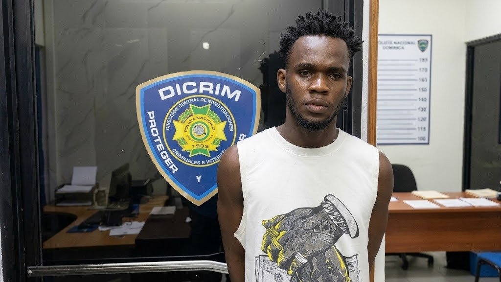 Un hombre joven con camiseta blanca sin mangas posa frente a una puerta de vidrio con el escudo de la Dirección Central de Investigaciones (Dicrim) de la Policía Nacional