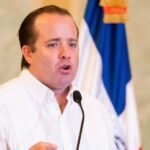 José Ignacio Paliza habla ante un micrófono con camisa blanca y una bandera dominicana detrás
