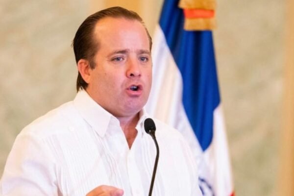 José Ignacio Paliza habla ante un micrófono con camisa blanca y una bandera dominicana detrás
