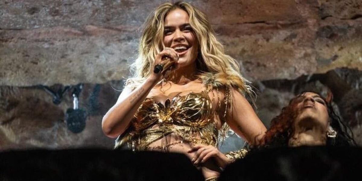 Karol G sonríe y canta con un micrófono durante un concierto, vestida con un corpiño dorado, junto a una bailarina