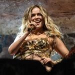 Karol G sonríe y canta con un micrófono durante un concierto, vestida con un corpiño dorado, junto a una bailarina