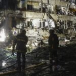 Dos bomberos de noche frente a un edificio residencial con fachada destruida y escombros