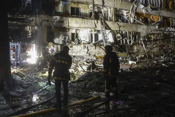 Dos bomberos de noche frente a un edificio residencial con fachada destruida y escombros
