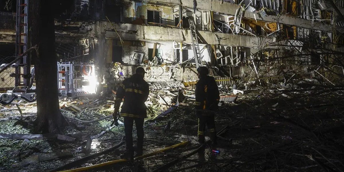 Dos bomberos de noche frente a un edificio residencial con fachada destruida y escombros