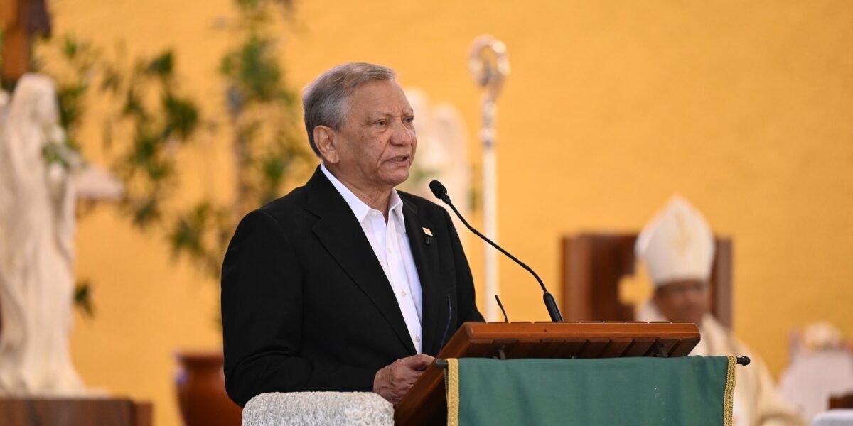 Un hombre de cabello canoso, con saco negro y camisa blanca, habla en un atril de una iglesia mientras un obispo con mitra lo escucha al fondo